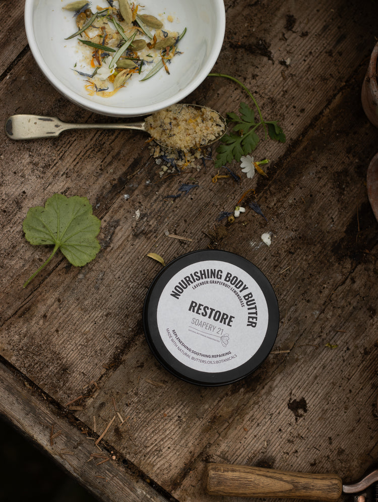 A bowl of botanical bath soaks sits on a rustic wooden table next to a tin of natural plant based body butter. there are leaves and other botanicals sprinkled on the table