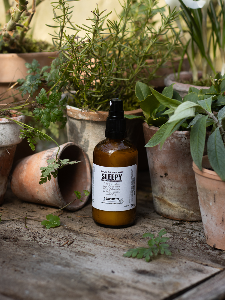 A bottle of natural aromatherapy room and linen mist named SLEEPY sits on a workbench surrounded by herbs in pots, the scene is natural and rustic