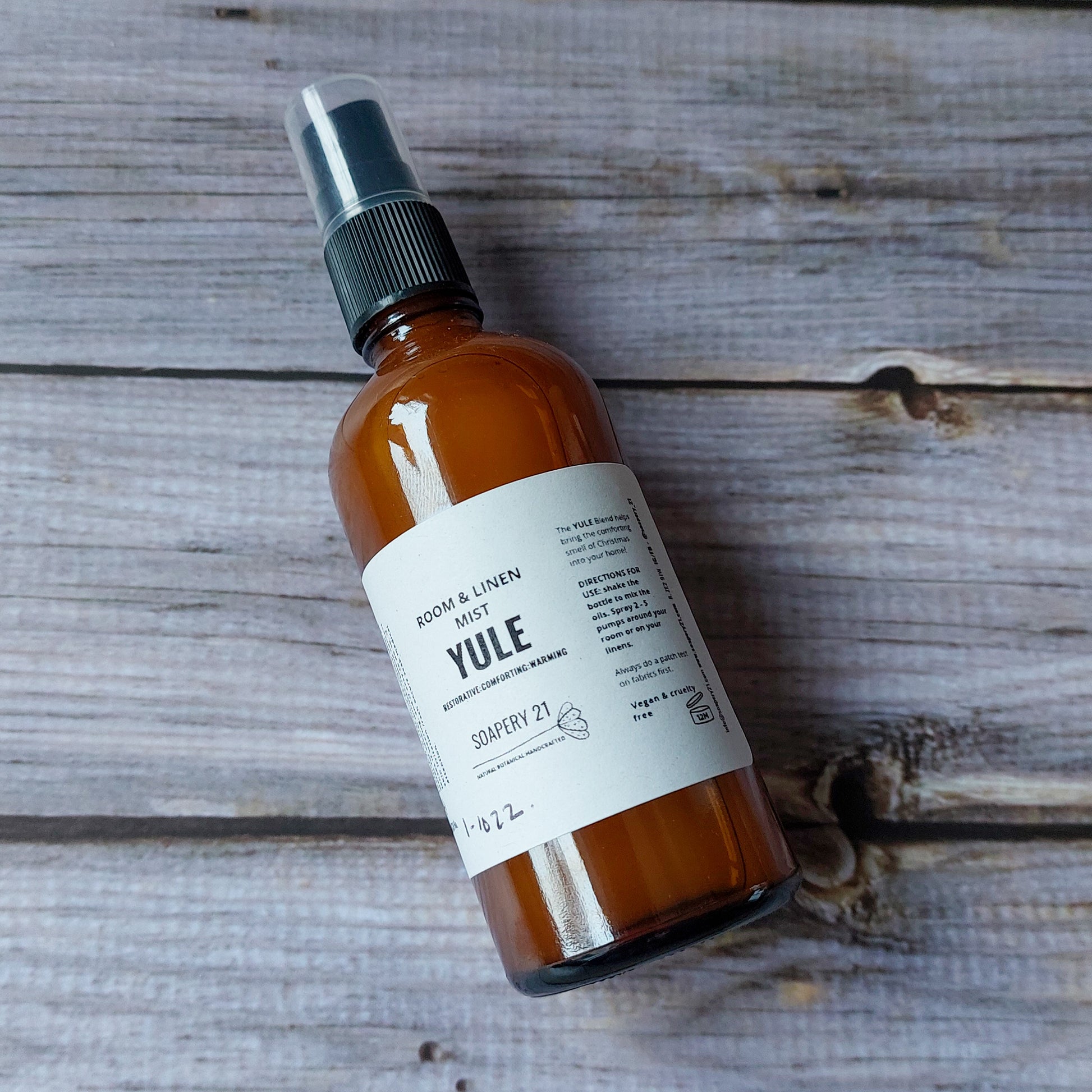 A brown glass bottle labeled "Yule Room &amp; Linen Mist" standing on a white wooden surface, surrounded by pinecones, a dried orange slice, and a cinnamon stick.